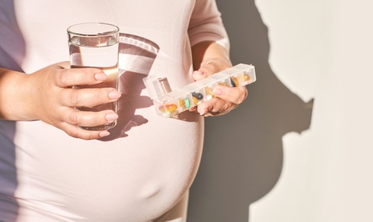 a woman holding a glass of water and a pill organizer in front of her preganant belly
